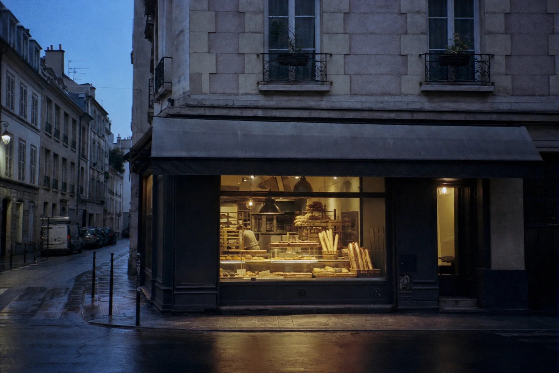 Devanture d'une petite boulangerie de quartier au petit matin, lumière chaude débordant sur le trottoir mouillé.